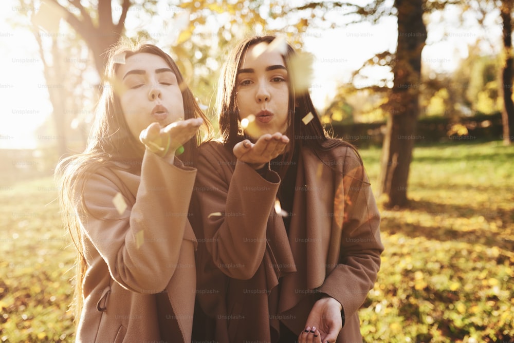Young brunette twin sisters standing close to each other and blowing confetti into camera, wearing casual coat in autumn sunny park on blurry background.
