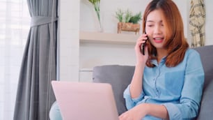 Business freelance Asian woman working on laptop and talking on phone with customer while sitting on table in living room. Lifestyle women working at home concept.