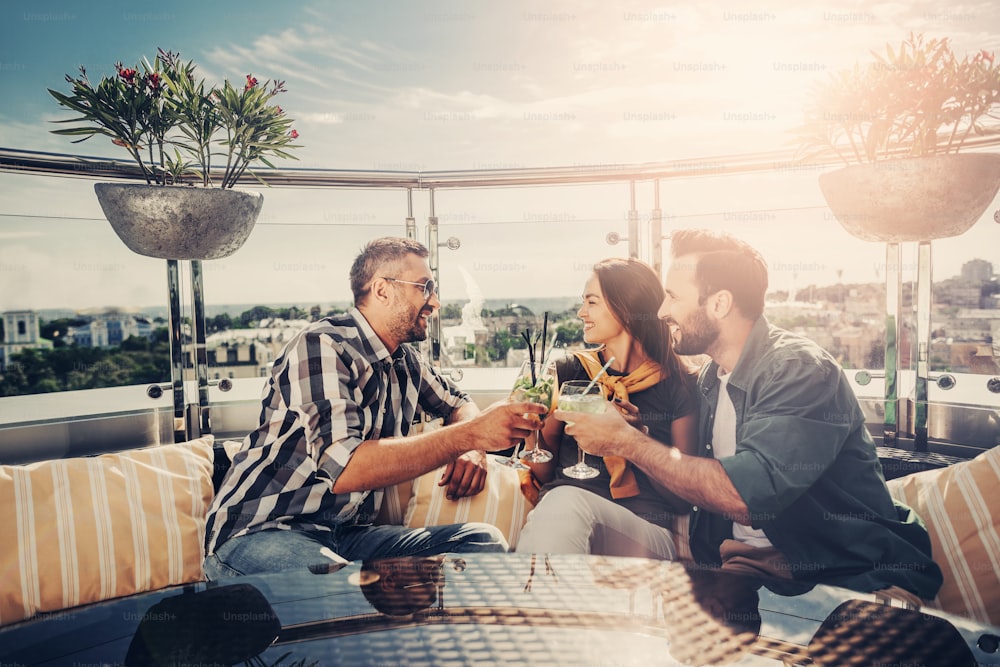 Feeling joyful. Bearded men and lovely young woman sitting at the table with alcohol drinks in their hands. They are toasting and laughing