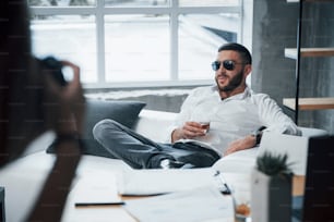 Dreamy atmosphere. Photograph on the left side. Young short-haired man in sunglasses sitting on couch in the office.