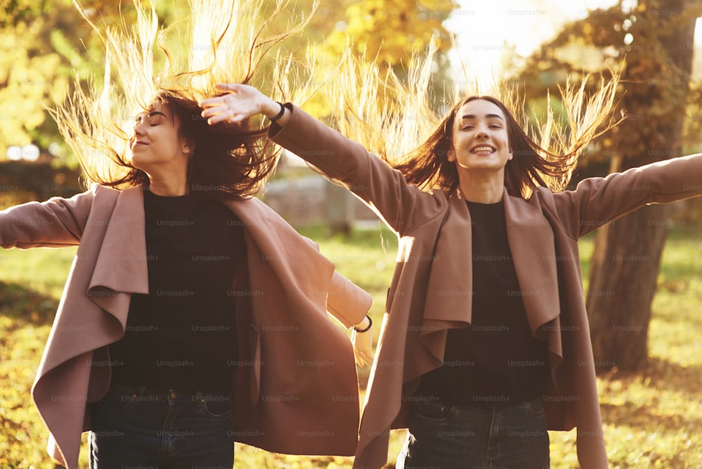 Young happy, smiling brunette twin girls standing and spreading hands wide open, their hair is dispelled up, wearing casual coat at autumn sunny park on the light blurry background. Freedom concept.