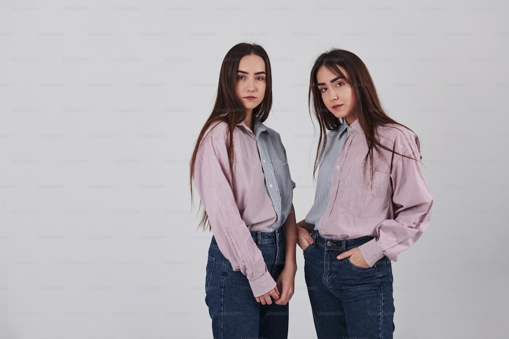 두 자매 쌍둥이가 흰색 배경의 스튜디오에 서서 포즈를 취하고 있습니다.