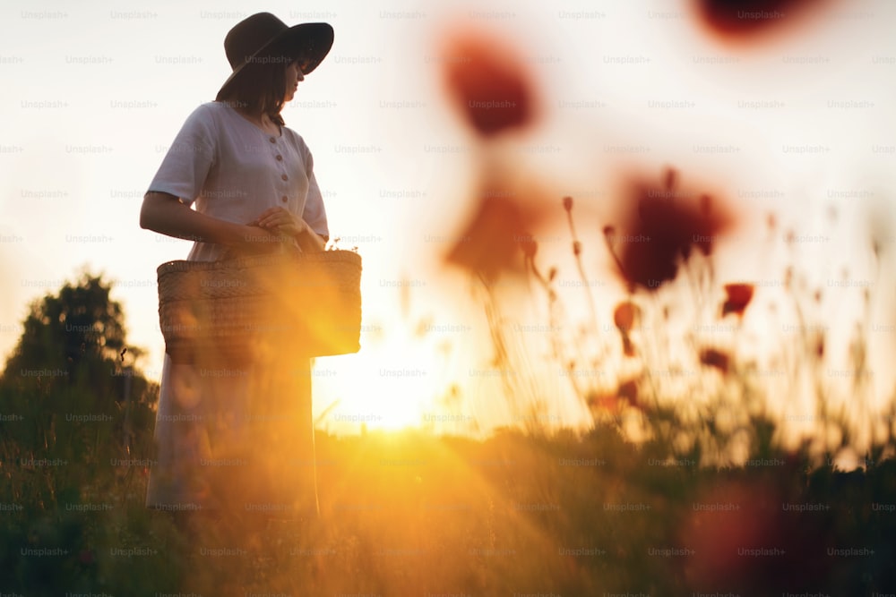 Fille élégante en robe de lin cueillant des fleurs dans un panier de paille rustique, marchant dans une prairie de coquelicots au coucher du soleil. Femme bohème au chapeau se relaxant dans la lumière chaude du soleil du soir dans le champ d’été