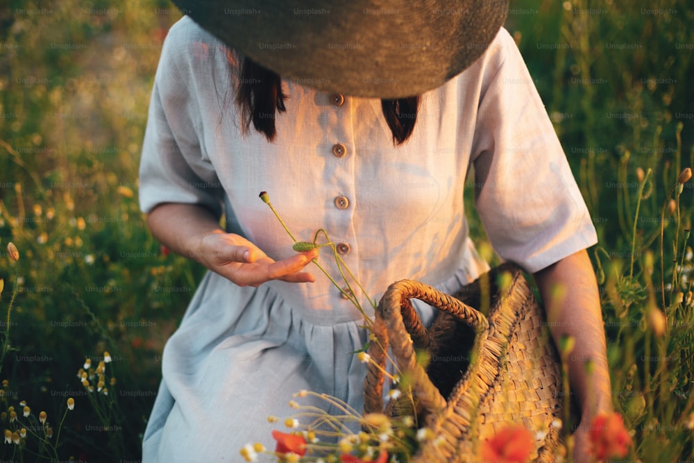 Stylish girl in linen dress gathering flowers in rustic straw basket, sitting in poppy meadow in sunset. Boho woman holding wildflowers bud in warm sunlight in summer field. Atmospheric