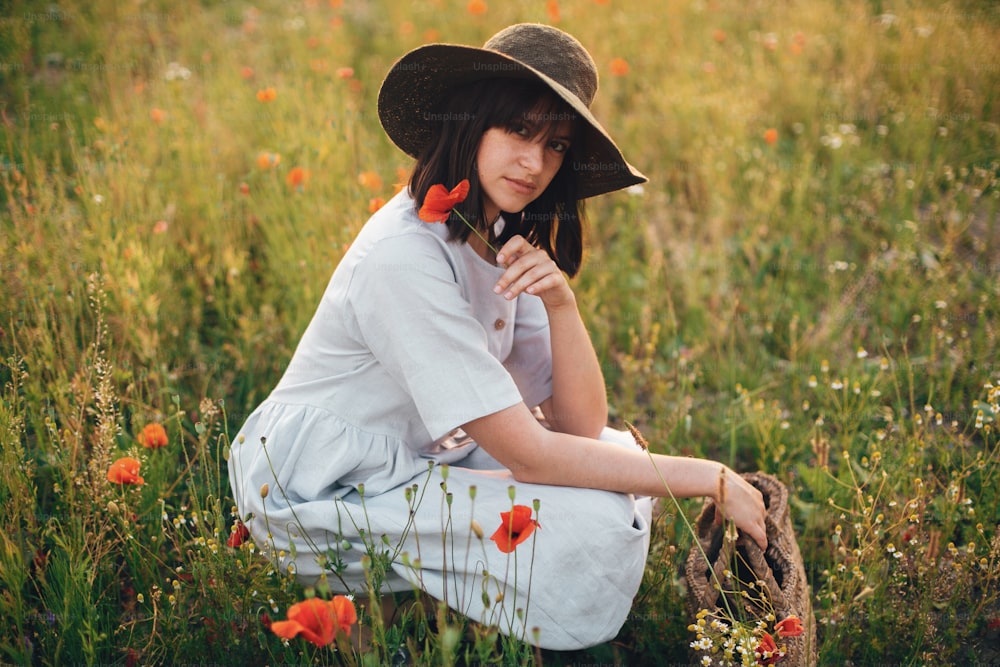 Menina elegante no vestido de linho segurando a flor da papoula no prado na luz do pôr do sol com flores na cesta de palha rústica. Mulher de Boho no chapéu relaxando no campo de verão. Momento atmosférico