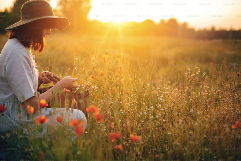 Muchacha elegante en vestido de lino recogiendo flores en cesta de paja rústica, sentada en prado de amapola al atardecer. Mujer boho en sombrero relajándose en la cálida luz del sol de la tarde en el campo de verano. Espacio para el texto