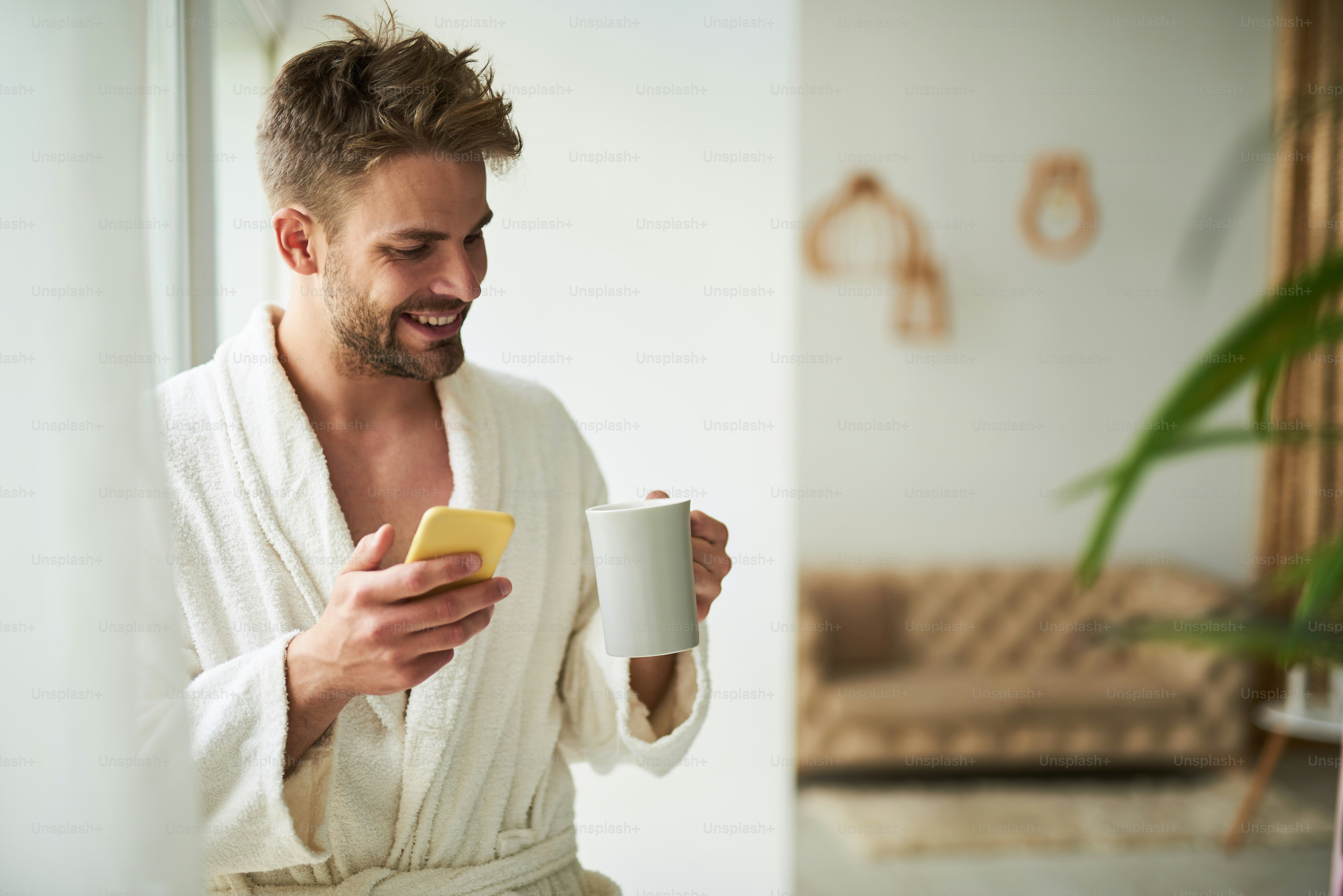朝、電話でおしゃべりする陽気な男性の横顔。白いバスローブを着て、コーヒーマグを片手に窓辺に立って微笑んでいる。右側のスペースをコピー