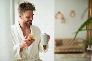 Side view of cheerful male chatting on phone in morning. He is standing by window in white bathrobe with coffee mug in hand and smiling. Copy space in right side