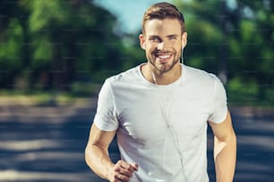 Mode de vie actif. Portrait d’un jeune homme positif profite d’un jogging matinal tout en écoutant de la musique dans des écouteurs. Il regarde la caméra avec le sourire tout en profitant de la chaude journée