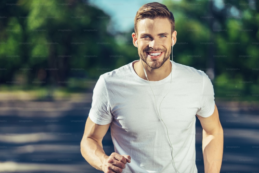 Active lifestyle. Portrait of positive young man is enjoying morning jogging while listening to music through earphones. He is looking at camera with smile while having pleasure from warm day