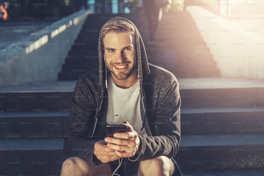 Portrait of young delighted gorgeous guy is sitting on stairs while spending time outdoors. He is looking at camera with joy while holding smartphone and listening to music through earphones