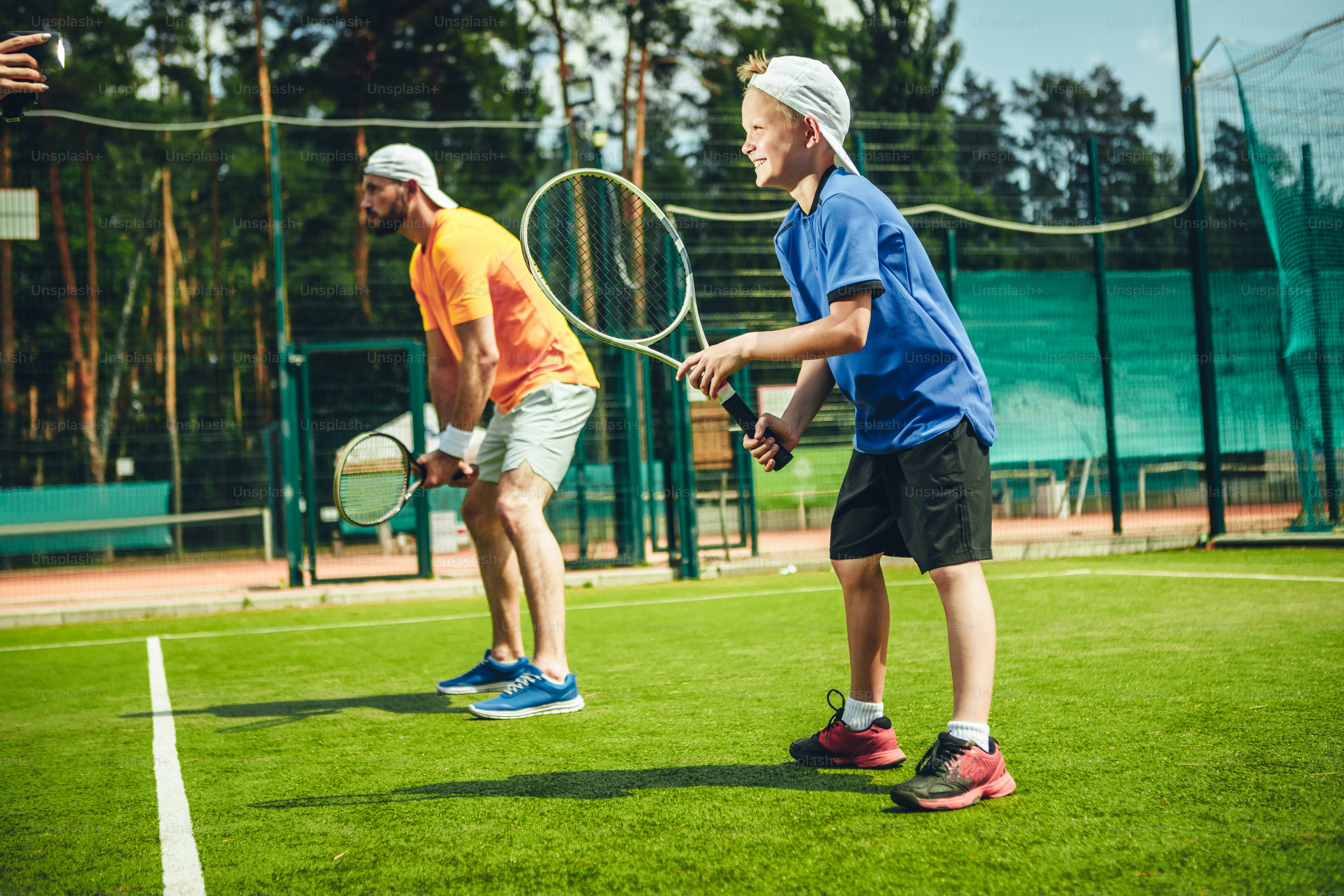 Full length side view happy man and beaming child playing tennis while situating on modern field with green grass