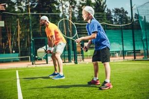Vue latérale pleine longueur homme heureux et enfant rayonnant jouant au tennis tout en se situant sur un terrain moderne avec de l’herbe verte