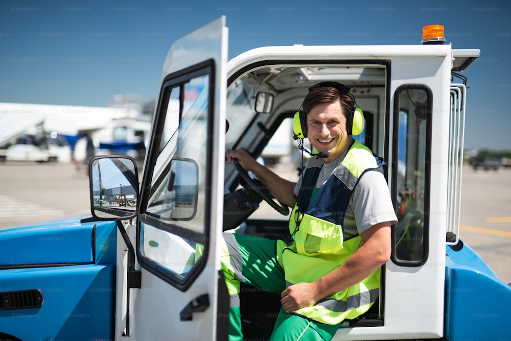 Joyful day. Cheerful man posing at camera before driving. He is holding the wheel and wearing headset