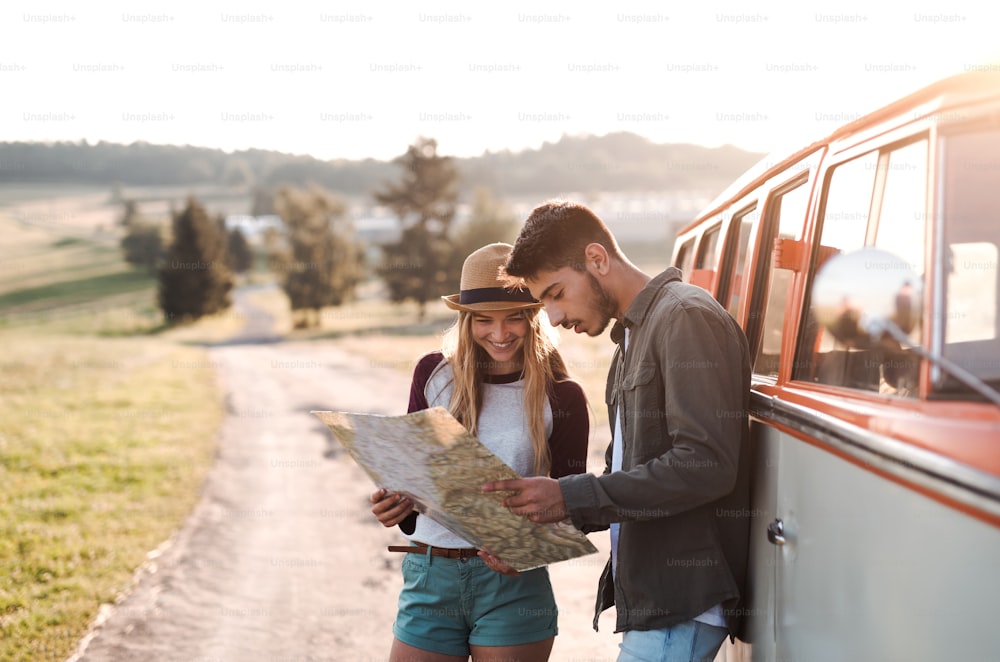 田舎をドライブトリップする若いカップルが、レトロなミニバンのそばに立って地図を見ている。