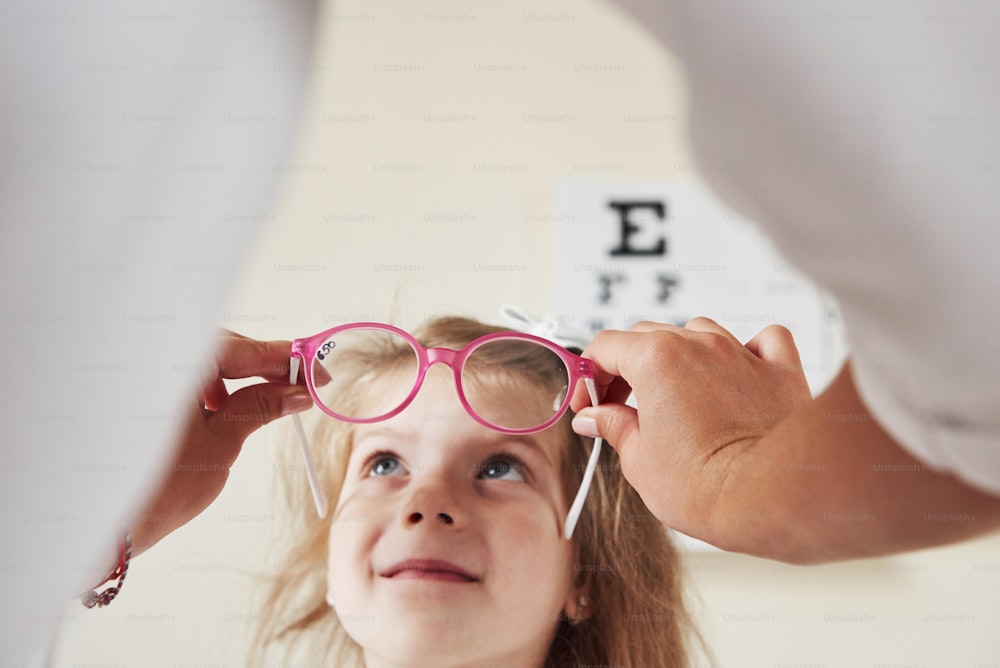 背景の視力をチェックするためのボードで新しいピンクのメガネを試している小さな女の子。
