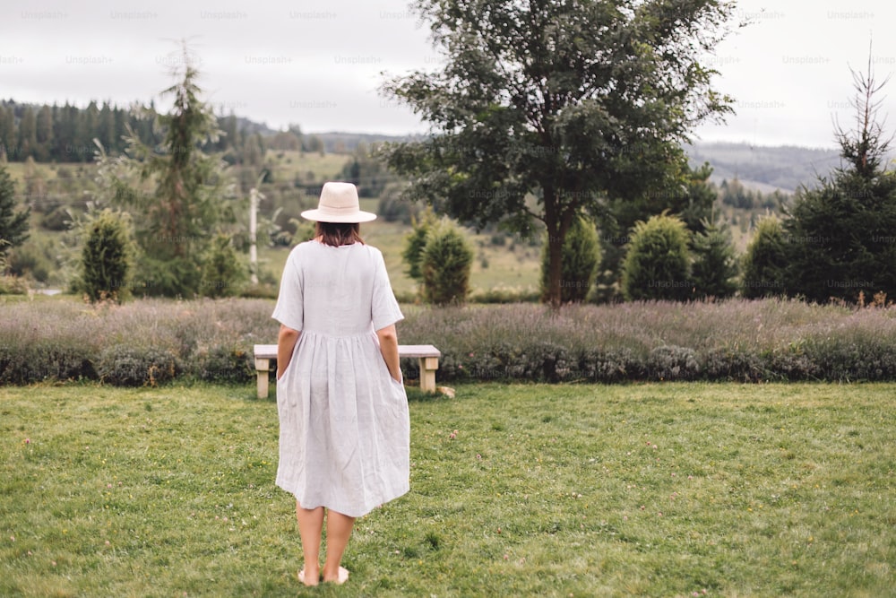 린넨 드레스와 모자를 쓴 세련된 힙스터 소녀가 라벤더 밭에 서서 산에서 휴식을 취하고 있습니다. 뒷모습. 보헤미안 여자는 웃으며 휴가를 즐기고 있다. 분위기 있는 소박한 순간. 복사 공간