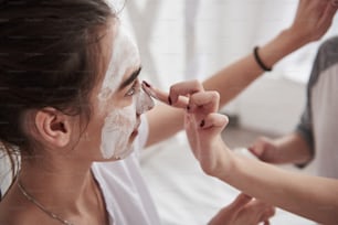 Close up view. Conception of skin care by using white mask on the face. Two female sisters have weekend at bedroom.