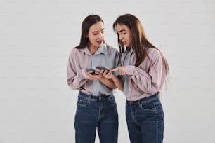 휴대 전화에서 흥미로운 것들을 보여줍니다. 두 자매 쌍둥이가 흰색 배경의 스튜디오에 서서 포즈를 취하고 있습니다.