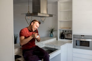 Young beautiful man having healthy breakfast while on the mobile phone at home in the kitchen
