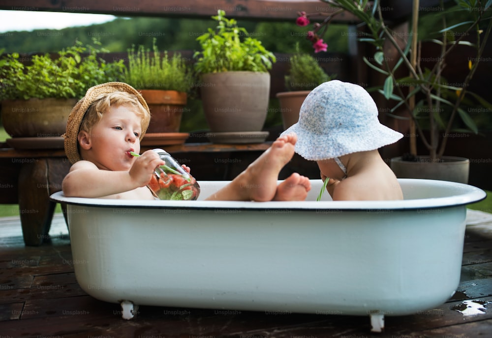 Bambini piccoli con una bevanda seduta in bagno all'aperto in giardino in estate, giocando in acqua.