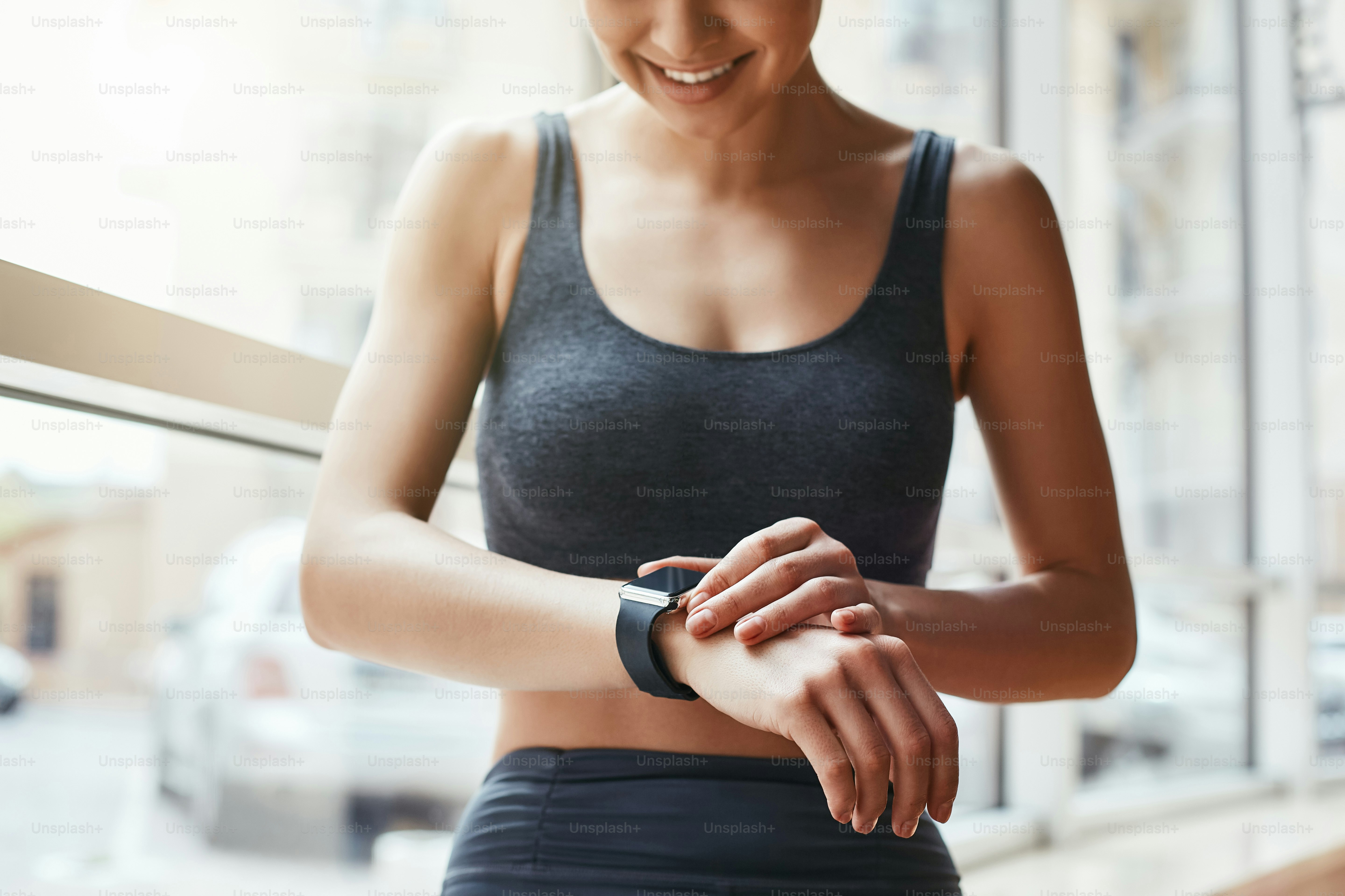 Checking all updates. Cropped photo of beautiful young woman looking at her sport bracelet while standing in front of window at gym. Fitness concept. Sport and healthy lifestyle. Technologies