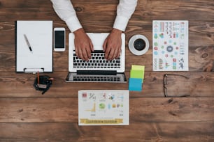 Top view of human hands typing on laptop. Laptop, smartphone, diary, coffee cup, glasses, charts and other supplies on brown wooden desk. Man working in his office. Horizontal shot