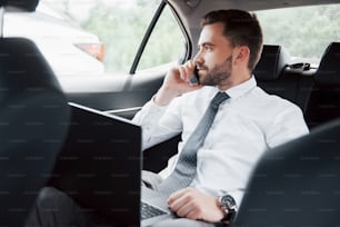 A young businessman working on laptop and talking on the phone while sitting in the car's back. Works in motion, appreciates its time.