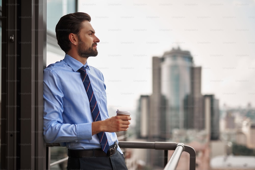 Corporate time-out culture. Waist up side on portrait of businessman leaning doorway of office balcony with cup of coffee and looking at city view