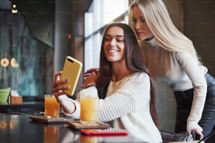 Let's take photo. In the restaurant. Two female friends sit indoors with yellow drink and use the smartphone.
