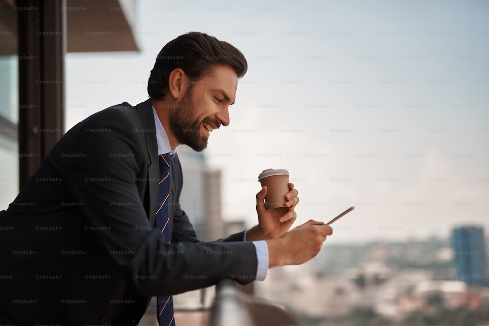 Take a pause. Waist up portrait of smiling businessman reading massages on smartphone while having coffee break on office balcony