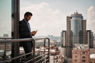 Take a pause. Full length portrait of smiling businessman reading massages on smartphone while standing on office terrace. Copy space on right