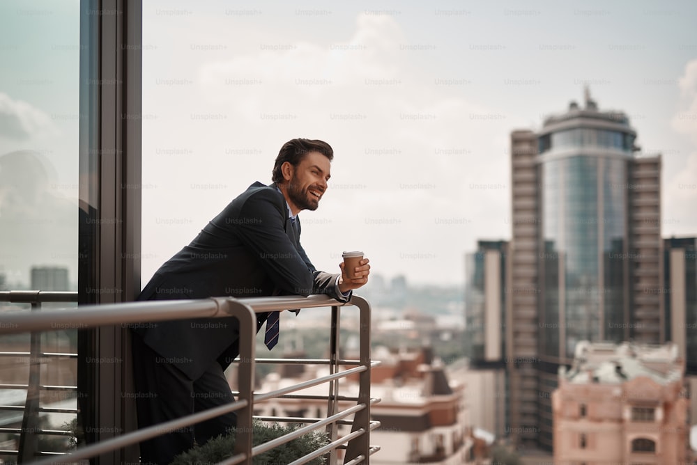 Take a pause. Full length portrait of cheerful businessman enjoying city view on office balcony with cup of coffee. Copy space on right