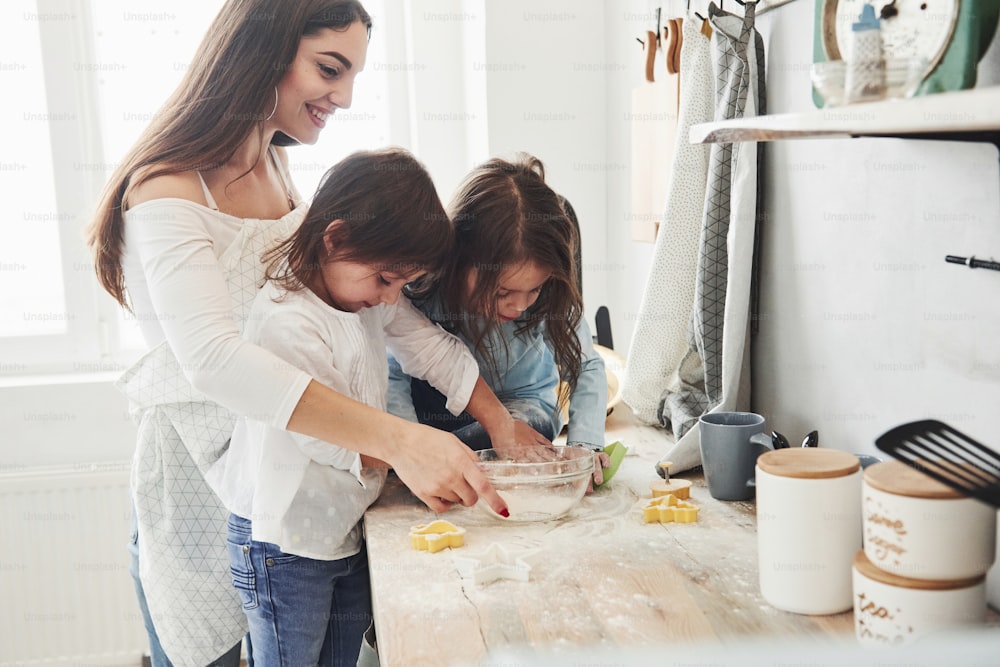 Vista lateral. Mamãe e duas meninas na cozinha está aprendendo a cozinhar uma boa comida com farinha.