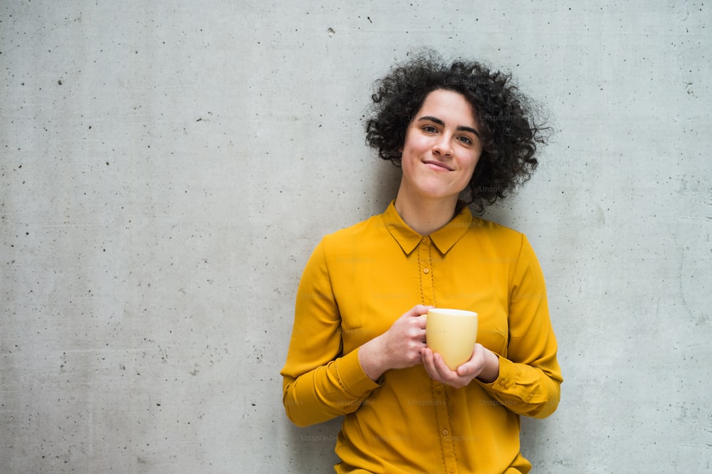 Ein Porträt einer jungen, fröhlichen Geschäftsfrau, die im Büro steht und eine Tasse Kaffee in der Hand hält.