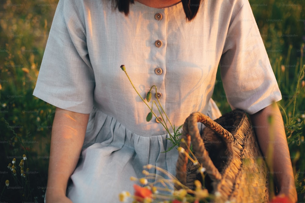 Fille élégante en robe de lin cueillant des fleurs dans un panier de paille rustique, assise dans une prairie de coquelicots au coucher du soleil. Femme bohème tenant un bourgeon de fleurs sauvages au soleil chaud dans un champ d’été.