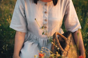 Stylish girl in linen dress gathering flowers in rustic straw basket, sitting in poppy meadow in sunset. Boho woman holding wildflowers bud in warm sunlight in summer field.