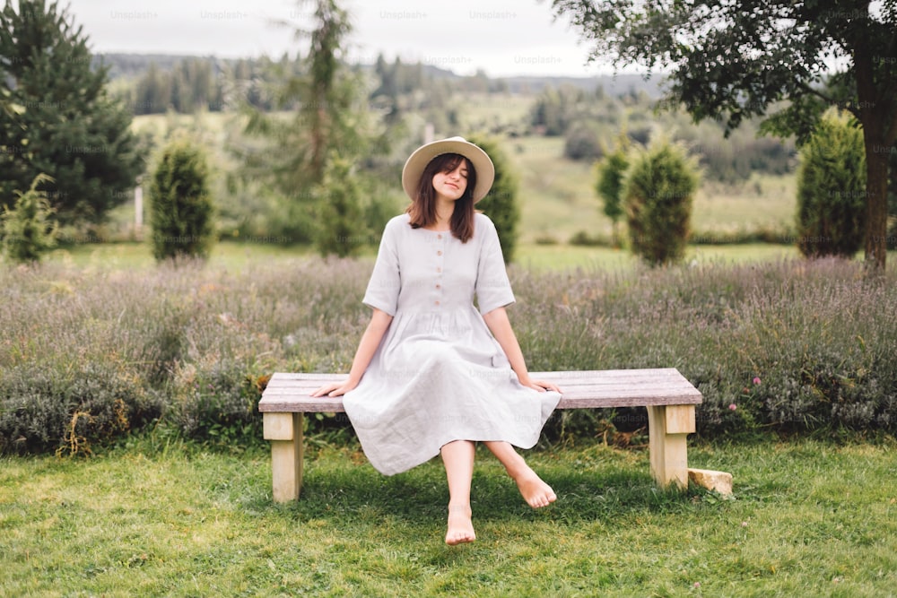 Stylish hipster girl in linen dress and hat sitting on bench at lavender field and relaxing in the morning. Happy bohemian woman enjoying vacation in mountains. Atmospheric rustic moment