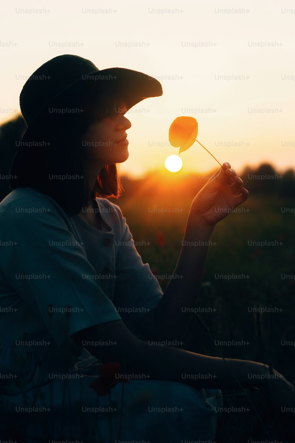 Silueta de una chica elegante en vestido de lino sosteniendo una flor de amapola en el prado a la luz del sol con flores en una cesta de paja rústica. Mujer boho en sombrero relajándose en el campo de la puesta del sol del verano. Atmosférico