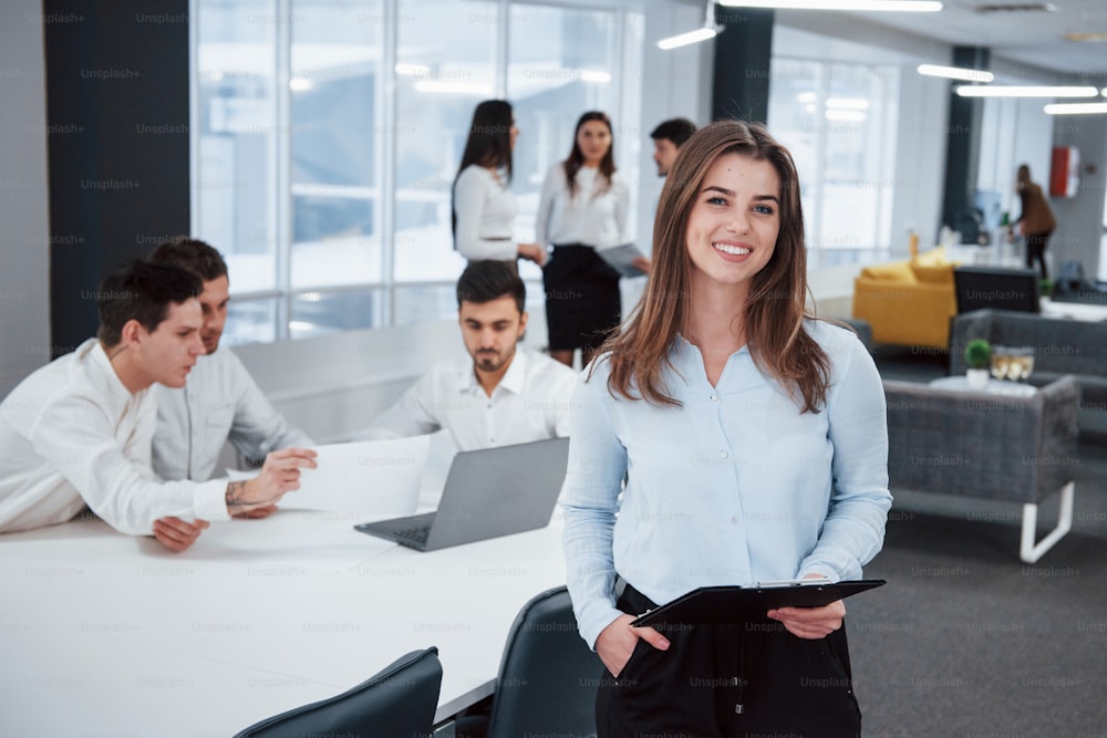 Relaxed woman indoors. Portrait of young girl stands in the office with employees at background.