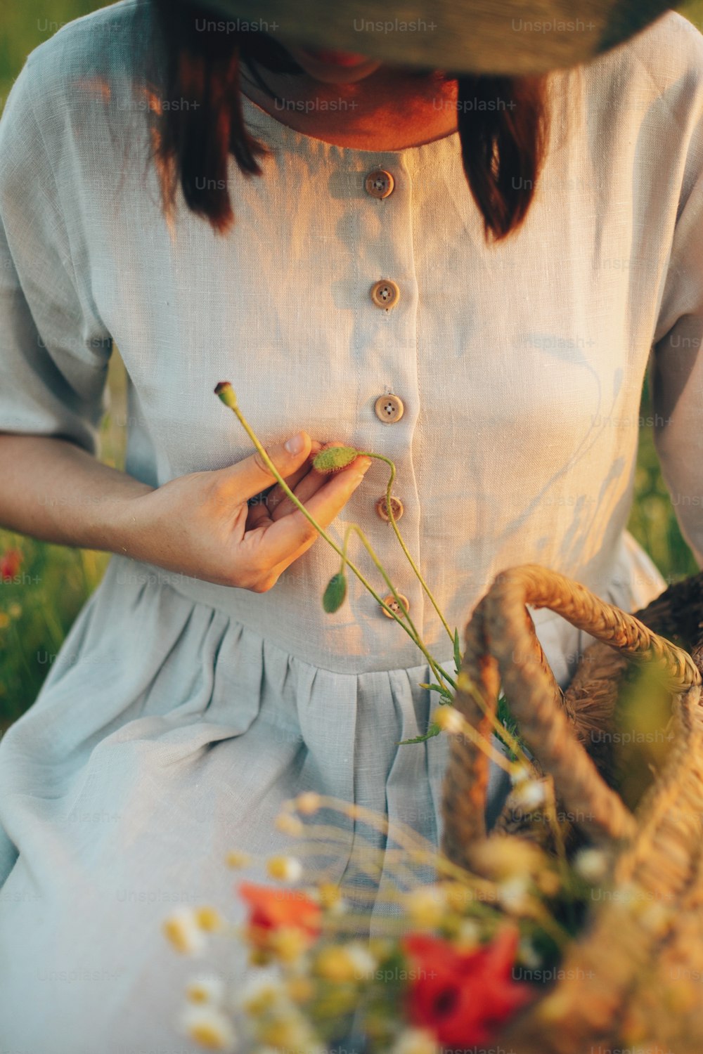 Muchacha elegante en vestido de lino recogiendo flores en cesta de paja rústica, sentada en prado de amapola al atardecer. Mujer boho sosteniendo capullos de flores silvestres a la cálida luz del sol en el campo de verano. Atmosférico