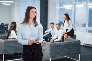 Smiling and looking to the side. Portrait of young girl stands in the office with employees at background.