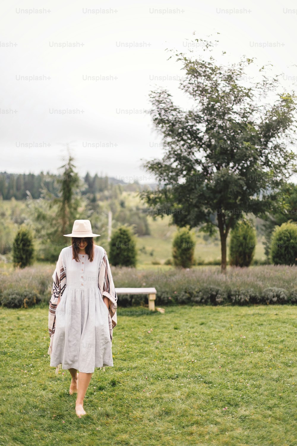 Bohemian woman smiling and enjoying vacation. Atmospheric rustic moment. Copy space. Stylish hipster girl in linen dress and hat walking at lavender field and relaxing in mountains