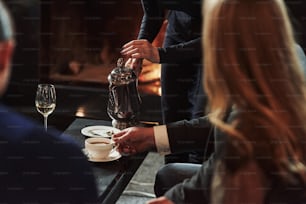 Cameriere femminile che versa tè caldo nella tazza bianca. Gli amici si siedono nel ristorante con un bellissimo camino.