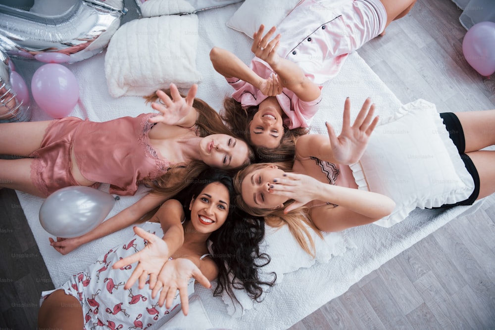 These beautiful friends wish you happy holidays. Top view of young girls at bachelorette party lying on the sofa and raising their hands up.