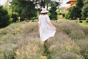 Stylish hipster girl in linen dress and hat walking in lavender field and relaxing. Happy bohemian woman enjoying lavender aroma. Back view. Atmospheric calm rural moment. Space for text