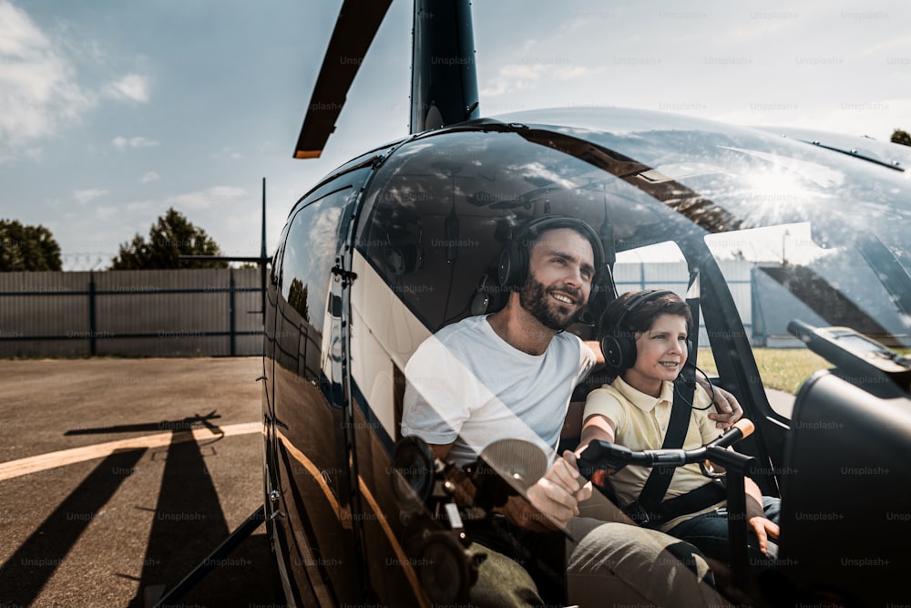 Portrait of happy unshaven father embracing glad boy while controlling helicopter with him. They sitting in cabin of it while talking. Optimistic parent and guy wearing earphones with microphones during sunny day