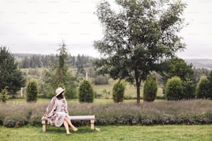 Chica hipster con estilo en vestido de lino y sombrero sentada en el banco en el campo de lavanda y relajándose por la mañana. Mujer bohemia feliz disfrutando de vacaciones en las montañas. Momento rústico atmosférico