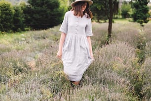 Chica hipster con estilo en vestido de lino y sombrero caminando en el campo de lavanda y sonriendo. Mujer bohemia feliz relajándose y disfrutando del aroma de lavanda. Momento rural de calma atmosférica. Espacio para el texto