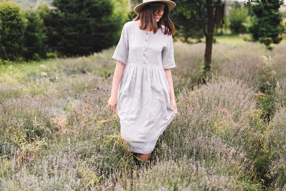 Stylish hipster girl in linen dress and hat walking in lavender field and smiling. Happy bohemian woman relaxing and enjoying lavender aroma. Atmospheric calm rural moment. Space for text